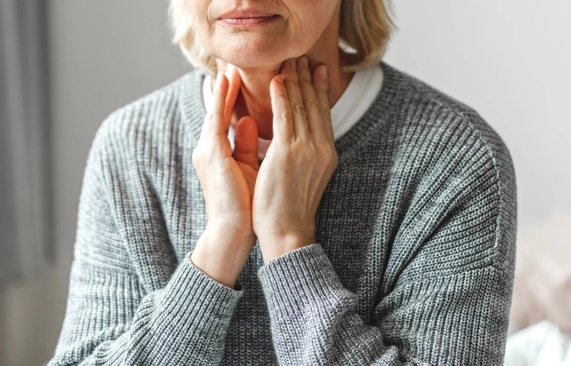 An older woman in a grey sweater touches her neck.